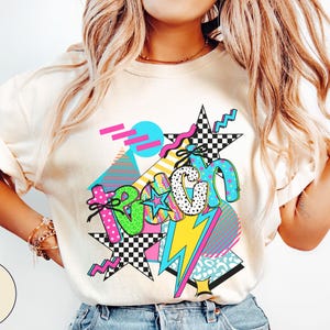 Puede incluir: Camiseta de color marfil con un dise&ntilde;o gr&aacute;fico colorido. El dise&ntilde;o presenta la palabra "Beach" en una fuente retro, con estrellas, rayos y formas geom&eacute;tricas en rosa, verde, azul y amarillo. La camiseta es de estilo casual.