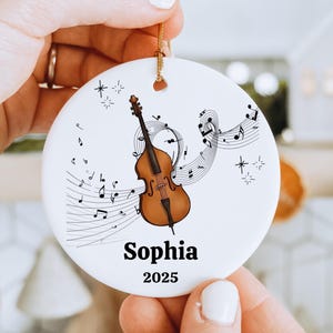 May include: A white ceramic ornament featuring a brown double bass and musical notes. The ornament has the name "Sophia" and the year "2025" printed in black. A gold string is attached for hanging. The ornament is held by a person.