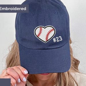 May include: Navy blue baseball cap with an embroidered baseball heart design. The heart is white with red stitching, and the number 23 is embroidered in white. The word "Embroidered" is above the cap.