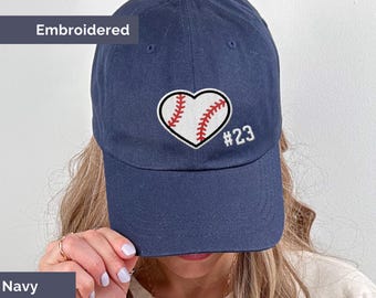 May include: Navy blue baseball cap with an embroidered baseball heart design. The heart is white with red stitching, and the number 23 is embroidered in white. The word "Embroidered" is above the cap.