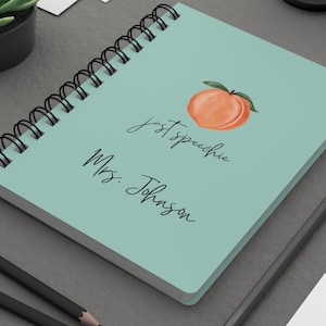 May include: A spiral-bound notebook with a light teal cover featuring a watercolor peach illustration and the text "just speechie Mrs. Johnson" in black script. A pencil and succulent are visible.