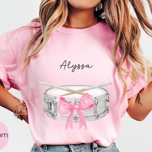 Puede incluir: Camiseta rosa claro con una ilustración de acuarela de una caja, baquetas y un lazo rosa. El nombre "Alyssa" está escrito encima del tambor en cursiva. La palabra "Blossom" está en un círculo rosa en la esquina inferior izquierda.