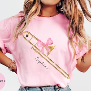 May include: A light pink t-shirt featuring a gold trombone design with a pink bow. The name "Sophia" is written in script below the instrument. The word "Blossom" is in a pink circle in the lower left corner.