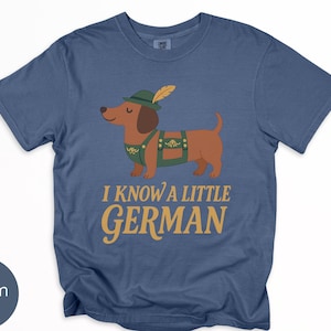 May include: Denim blue t-shirt featuring a cartoon dachshund wearing a green hat and lederhosen. The text "I KNOW A LITTLE GERMAN" is printed in gold. The shirt is a casual style.