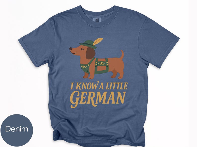 May include: Denim blue t-shirt featuring a cartoon dachshund wearing a green hat and lederhosen. The text "I KNOW A LITTLE GERMAN" is printed in gold. The shirt is a casual style.