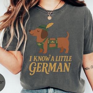 May include: Dark gray t-shirt featuring a cartoon dachshund wearing a green hat and lederhosen. The text "I KNOW A LITTLE GERMAN" is printed in gold. The shirt is a comfortable, casual style.