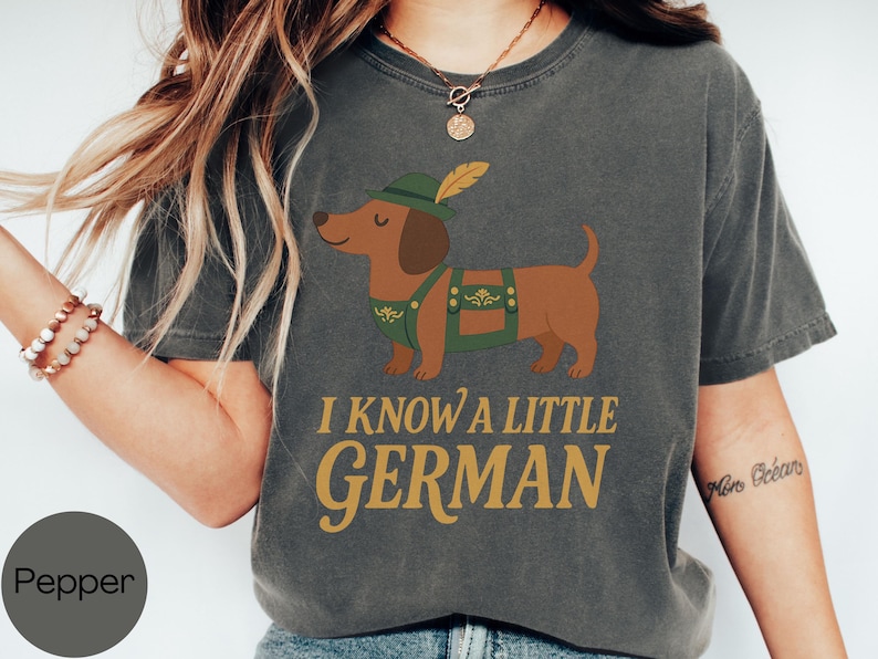 May include: Dark gray t-shirt featuring a cartoon dachshund wearing a green hat and lederhosen. The text "I KNOW A LITTLE GERMAN" is printed in gold. The shirt is a comfortable, casual style.