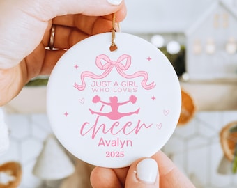 May include: A white ceramic ornament with a pink bow, ribbons, and text that reads "Just a girl who loves cheer" with a cheerleader graphic. The ornament also includes the name "Avalyn" and the year "2025".
