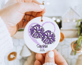 May include: White ceramic ornament featuring two purple and white pom-poms inside a purple heart outline. The name "Sophia" and the year "2025" are printed below the pom-poms. A gold string is attached for hanging.