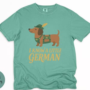 May include: Light green t-shirt with a cartoon dachshund wearing a green hat and lederhosen. The text "I KNOW A LITTLE GERMAN" is printed in gold letters below the dog. The shirt is made of a soft material.