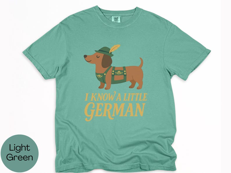 May include: Light green t-shirt with a cartoon dachshund wearing a green hat and lederhosen. The text "I KNOW A LITTLE GERMAN" is printed in gold letters below the dog. The shirt is made of a soft material.