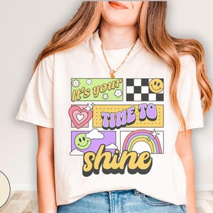 May include: Ivory t-shirt with a retro design. The design features the text "It's your time to shine" in a colourful, blocky font. The graphic includes a smiley face, a heart, a rainbow, and a checkered pattern.