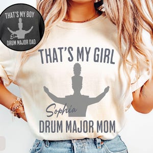 May include: Ivory-colored t-shirt with the text "THAT'S MY GIRL" and "DRUM MAJOR MOM" in gray. A silhouette of a drum major is featured below the text. A smaller image on the top left says "THAT'S MY BOY" and "DRUM MAJOR DAD".
