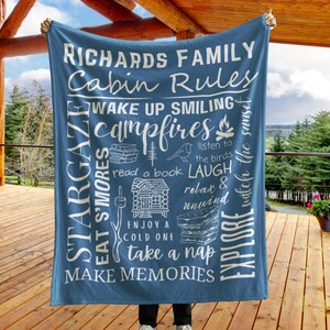 May include: A blue fleece blanket with white text that reads "Richards Family Cabin Rules" and includes a list of cabin rules such as "Wake Up Smiling", "Campfires", "Stargaze", "Eat S'mores", "Read a Book", "Laugh", "Relax & Unwind", "Enjoy a Cold One", "Take a Nap", "Explore", and "Make Memories".