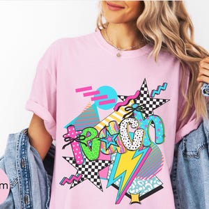 Puede incluir: Camiseta rosa con un dise&ntilde;o gr&aacute;fico colorido. El dise&ntilde;o presenta la palabra "TEACH" en varias fuentes y patrones, incluyendo lunares y estrellas a cuadros, con un rayo y formas geom&eacute;tricas. La camiseta se lleva con una chaqueta vaquera.