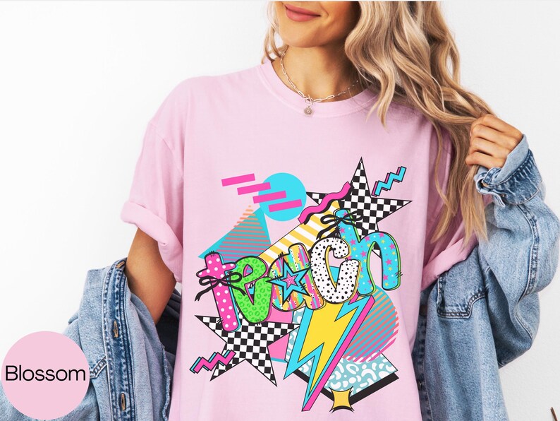Puede incluir: Camiseta rosa con un dise&ntilde;o gr&aacute;fico colorido. El dise&ntilde;o presenta la palabra "TEACH" en varias fuentes y patrones, incluyendo lunares y estrellas a cuadros, con un rayo y formas geom&eacute;tricas. La camiseta se lleva con una chaqueta vaquera.