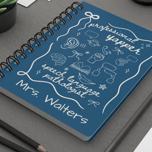May include: A blue spiral-bound notebook with white doodles and text. The notebook says "professional yapper" and "speech language pathologist Mrs. Walters". A pencil and a succulent plant are in the background.