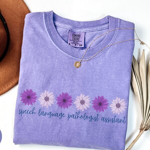 May include: A violet Comfort Colors t-shirt with the text "speech language pathologist assistant" in a script font. The shirt features a row of purple and white daisy graphics. A gold necklace and a brown hat are also visible.