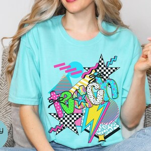 Puede incluir: Una camiseta azul claro con un dise&ntilde;o gr&aacute;fico colorido. El dise&ntilde;o incluye la palabra "Beach" en una mezcla de fuentes y patrones, junto con estrellas, un rayo y formas geom&eacute;tricas. La camiseta es de estilo casual.