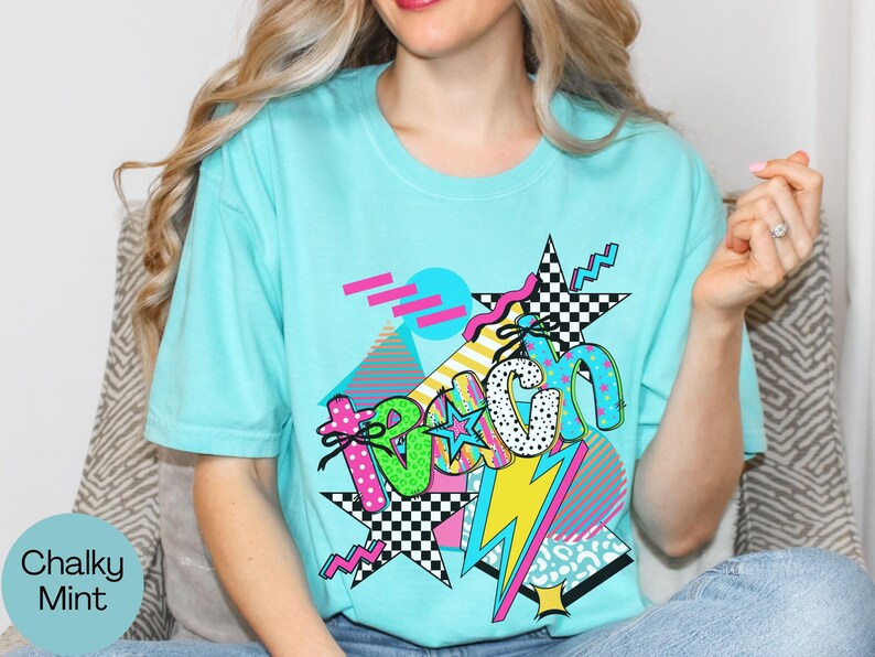 Puede incluir: Una camiseta azul claro con un dise&ntilde;o gr&aacute;fico colorido. El dise&ntilde;o incluye la palabra "Beach" en una mezcla de fuentes y patrones, junto con estrellas, un rayo y formas geom&eacute;tricas. La camiseta es de estilo casual.