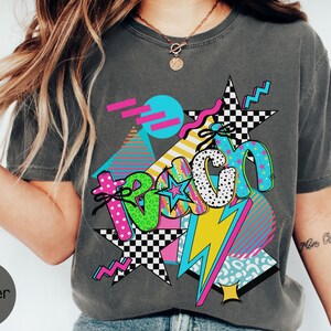 Puede incluir: Camiseta gris oscuro con un dise&ntilde;o gr&aacute;fico colorido. El dise&ntilde;o presenta la palabra "Teach" en una fuente l&uacute;dica, con estrellas, rayos y formas geom&eacute;tricas en colores vibrantes como rosa, azul, amarillo y verde. La camiseta tiene una est&eacute;tica vintage y retro.