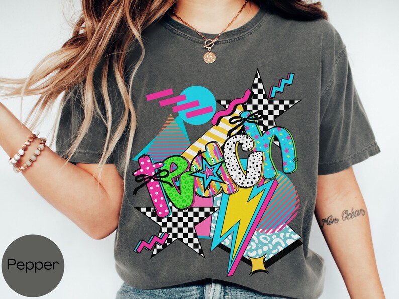 Puede incluir: Camiseta gris oscuro con un dise&ntilde;o gr&aacute;fico colorido. El dise&ntilde;o presenta la palabra "Teach" en una fuente l&uacute;dica, con estrellas, rayos y formas geom&eacute;tricas en colores vibrantes como rosa, azul, amarillo y verde. La camiseta tiene una est&eacute;tica vintage y retro.