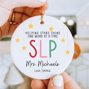 Peut inclure: Un ornement en céramique blanc avec une ficelle dorée, portant l'inscription "HELPING STARS SHINE ONE WORD AT A TIME" et "SLP" en rouge, rose et vert. L'ornement comprend également le nom "Mrs. Michaels" et le message "Love, Emma", avec des étoiles jaunes.