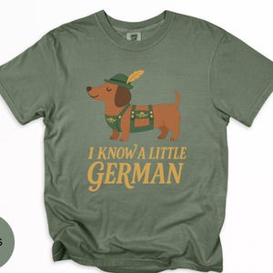 May include: Moss green t-shirt featuring a cartoon dachshund wearing a traditional German outfit, including a hat with a feather and lederhosen. The text "I KNOW A LITTLE GERMAN" is printed in gold lettering below the dog.