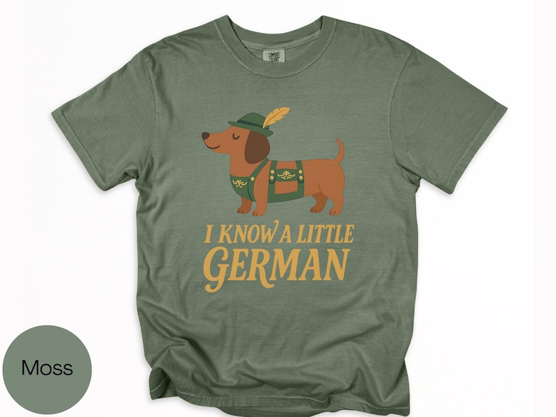 May include: Moss green t-shirt featuring a cartoon dachshund wearing a traditional German outfit, including a hat with a feather and lederhosen. The text "I KNOW A LITTLE GERMAN" is printed in gold lettering below the dog.
