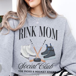 May include: A sport grey crewneck sweatshirt featuring the black text RINK MOM SOCIAL CLUB TOE PICKS & HOCKEY STICKS. The graphic includes a pair of white figure skates, a pair of black hockey skates, two crossed hockey sticks, and a black puck.