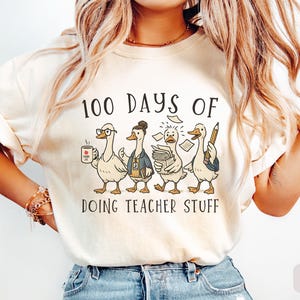 May include: Ivory t-shirt featuring the text "100 DAYS OF DOING TEACHER STUFF" and a graphic of four cartoon ducks dressed as teachers. One duck holds a coffee mug, another a stack of papers, and another a pencil.