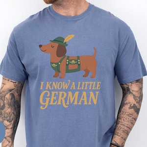 May include: Blue jean t-shirt featuring a cartoon dachshund wearing a green hat and lederhosen. The text "I KNOW A LITTLE GERMAN" is printed in gold. The dog is brown with a green and gold outfit.
