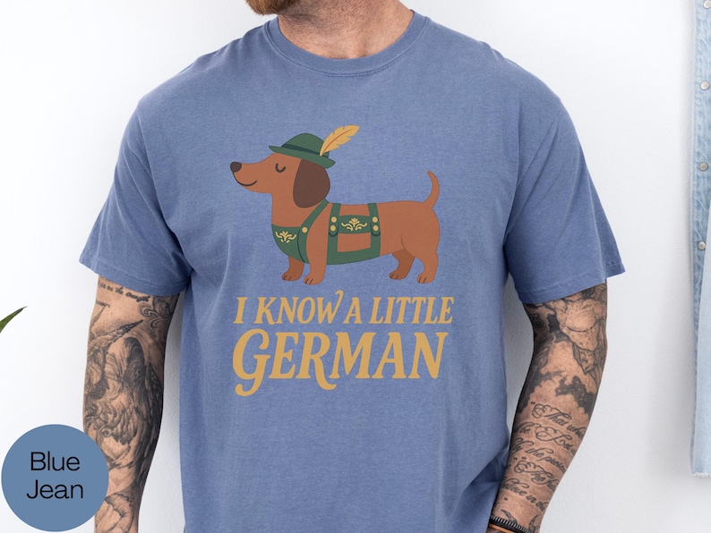 May include: Blue jean t-shirt featuring a cartoon dachshund wearing a green hat and lederhosen. The text "I KNOW A LITTLE GERMAN" is printed in gold. The dog is brown with a green and gold outfit.