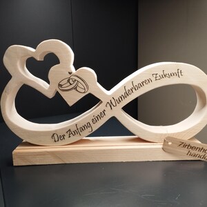 Può includere: Un simbolo di infinito in legno con due cuori intrecciati e due anelli all'interno di uno dei cuori. Il simbolo di infinito è inciso con il testo "Der Anfang einer wunderbaren Zukunft", che si traduce in "L'inizio di un futuro meraviglioso". La base in legno ha un piccolo cartellino che recita "Zirbenholz handgefertigt", che si traduce in "Legno di pino fatto a mano".