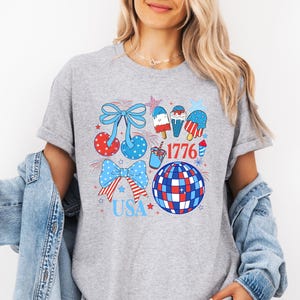 Puede incluir: Camiseta gris con un dise&ntilde;o patri&oacute;tico que presenta lazos, helados, una bola de discoteca y los textos "1776" y "USA" en rojo, blanco y azul. La camiseta tiene cuello redondo y mangas cortas.