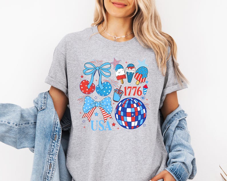 Puede incluir: Camiseta gris con un dise&ntilde;o patri&oacute;tico que presenta lazos, helados, una bola de discoteca y los textos "1776" y "USA" en rojo, blanco y azul. La camiseta tiene cuello redondo y mangas cortas.