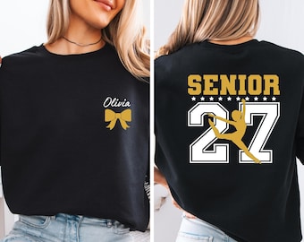 May include: A black crewneck sweatshirt featuring a gold bow and the name Olivia in white script on the front left chest. The back displays the word SENIOR in gold, a row of white stars, and the number 27 with a gold dancer silhouette.