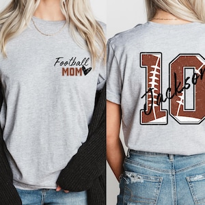 May include: Two gray t-shirts with a football theme. One shirt says "Football Mom" with a heart. The other shirt has a brown football jersey number "10" with the name "Jackson" written on it.