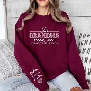 May include: Burgundy sweatshirt with the text "This Grandma wears her heart on her sleeve" and the names "Calvin, Karissa, Jessica" on the sleeve.