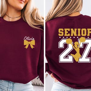 May include: A maroon crewneck sweatshirt featuring a gold bow and the name Olivia on the front left chest. The back displays the word SENIOR in gold above the number 27 in white, with a gold silhouette of a cheerleader holding pom-poms.