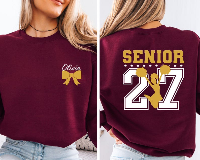 May include: A maroon crewneck sweatshirt featuring a gold bow and the name Olivia on the front left chest. The back displays the word SENIOR in gold above the number 27 in white, with a gold silhouette of a cheerleader holding pom-poms.
