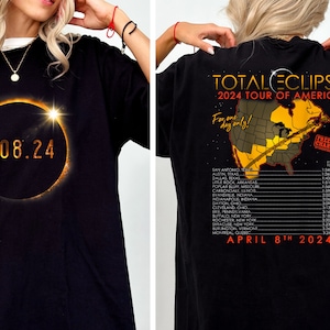 May include: Black t-shirt with a graphic of a solar eclipse and the date 4.08.24. The back of the shirt features a map of the United States with the text "TOTALECLIPSE 2024 TOUR OF AMERICA" and "For one day only!"  The text "FREE OF CHARGE" is in a red box on the map. A list of cities and times for the eclipse is printed below the map.