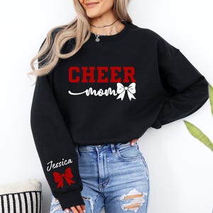 May include: Black sweatshirt with the text "CHEER mom" in red and white with a red and white bow. The sweatshirt has a white text "Jessica" on the left sleeve with a red and white bow.