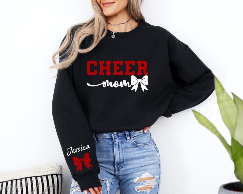 May include: Black sweatshirt with the text "CHEER mom" in red and white with a red and white bow. The sweatshirt has a white text "Jessica" on the left sleeve with a red and white bow.