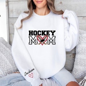 Pu&ograve; includere: Felpa bianca con il testo "HOCKEY MOM" in nero con un cuore rosso e bastoni da hockey. Il testo "Jackson" &egrave; scritto sulla manica con un cuore rosso.
