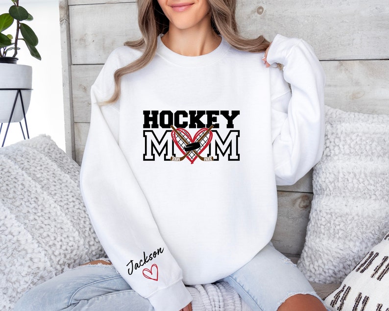 Pu&ograve; includere: Felpa bianca con il testo "HOCKEY MOM" in nero con un cuore rosso e bastoni da hockey. Il testo "Jackson" &egrave; scritto sulla manica con un cuore rosso.