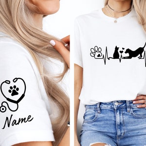 May include: White t-shirt with a black paw print and stethoscope design on the left sleeve. The right side of the shirt features a black paw print, cat, and dog with a heartbeat line and the text "Name".