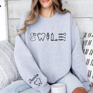 May include: A gray sweatshirt with the word "SMILE" written in black with dental icons. The left sleeve has the name "Jessica" written in black with a tooth icon.