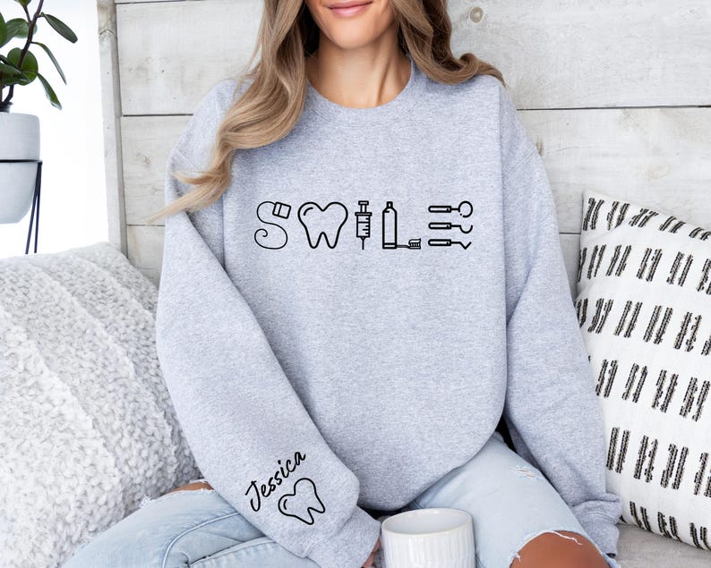 May include: A gray sweatshirt with the word "SMILE" written in black with dental icons. The left sleeve has the name "Jessica" written in black with a tooth icon.