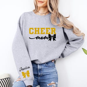 May include: Gray sweatshirt with the text "CHEER mom" in gold letters and a black bow. The left sleeve has the name "Jessica" in black letters and a gold bow.